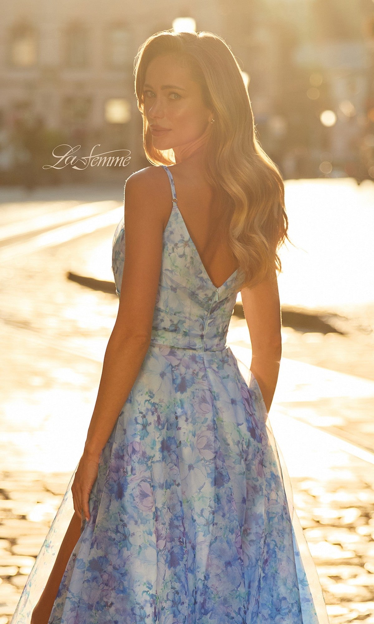 A woman stands outdoors in sunlight, looking over her shoulder, wearing the Long Prom Dress: La Femme 33502. The background is softly blurred with a warm glow.